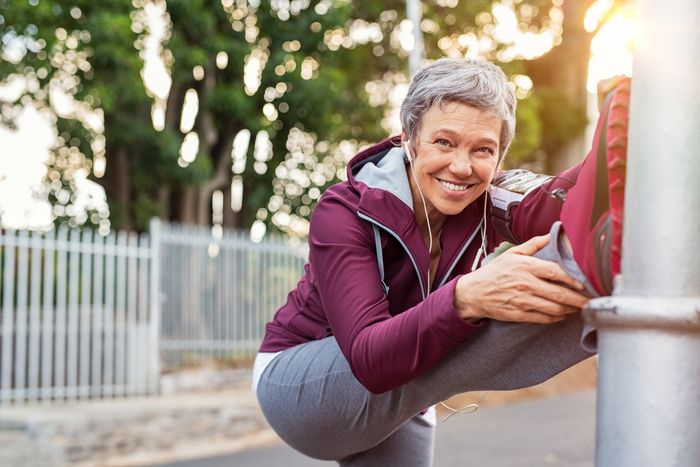 A woman wearing headphones and a jogging outfit smiles and stretches her leg.
